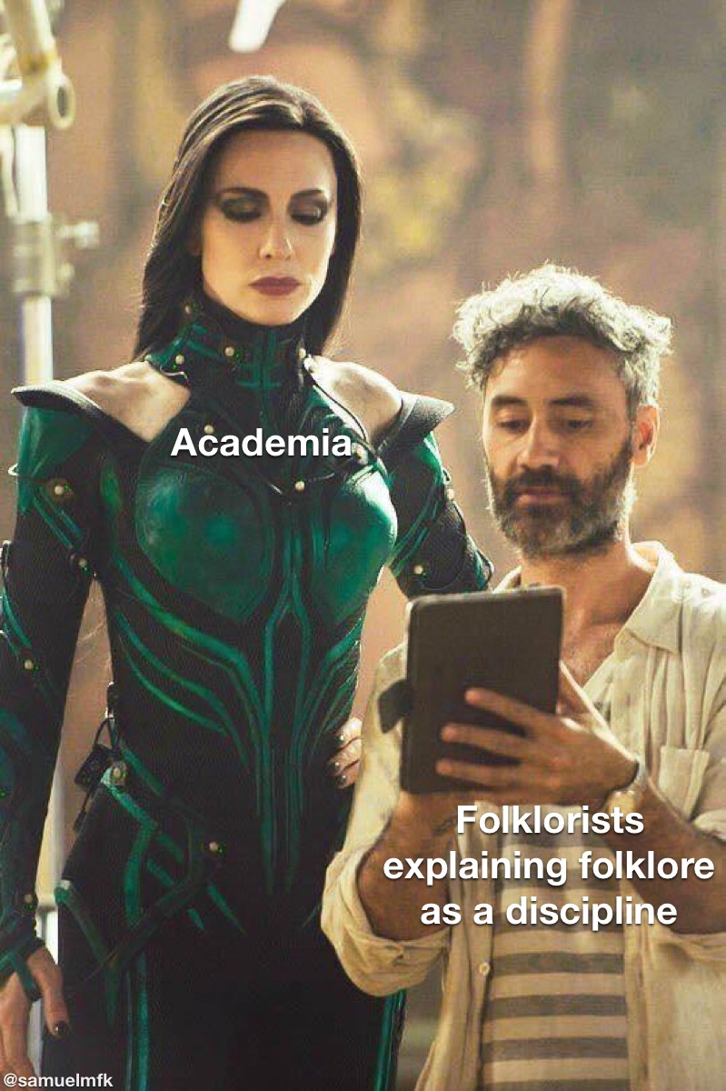 An image of film director Taika Waititi giving actress Cate Blanchett notes on the set of Thor: Ragnarok. Blanchett is wearing a green bodysuit and is standing over Waititi who appears to be reading from a series of notes. Blanchett is labeled “Academia” and Waititi is labeled “Folklorists explaining folklore as a discipline” in white text.